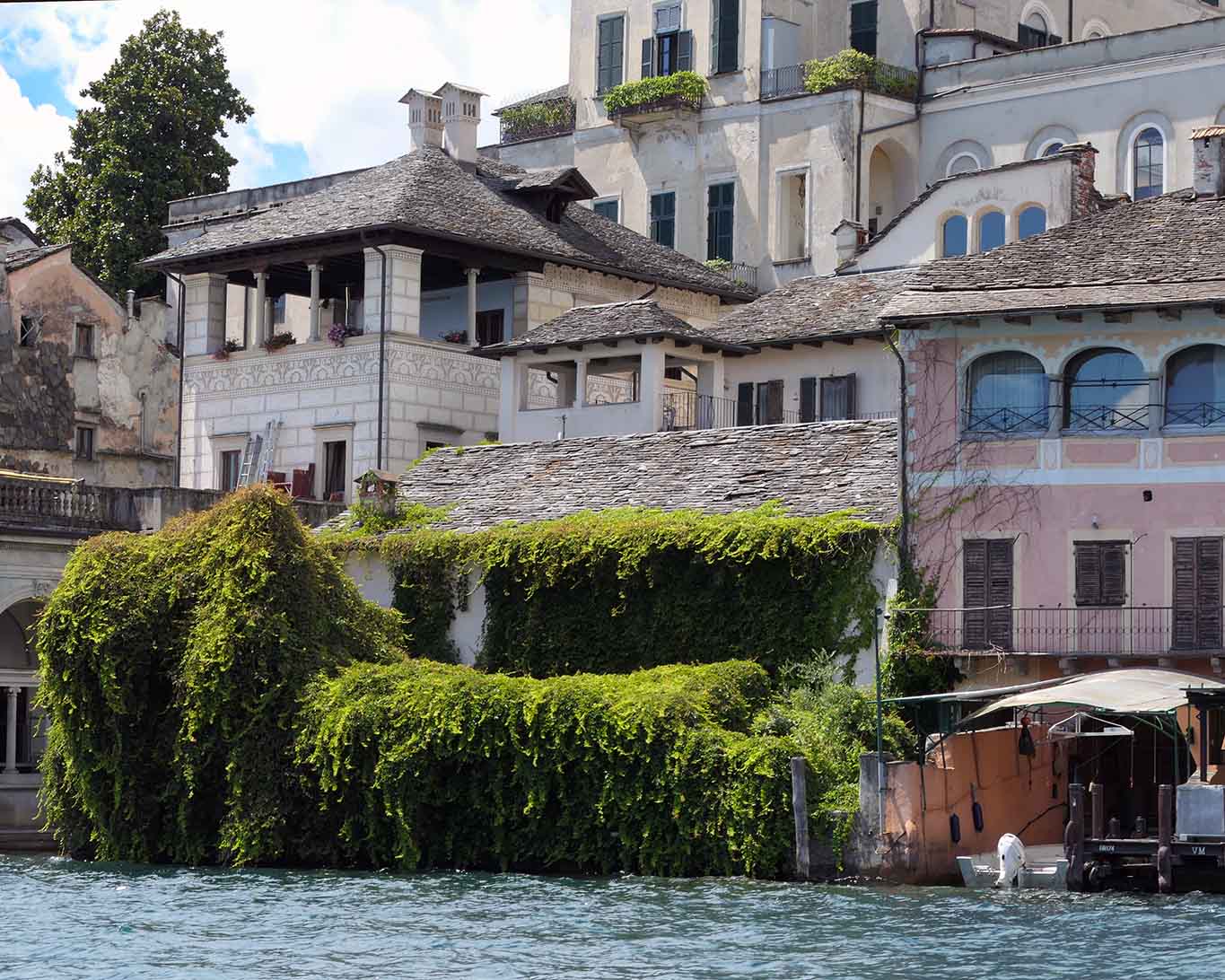 Üppiges Grün wuchert über altes Mauerwerk auf einer sonnenbeschienen Insel und Häusern mit schmucken Fassaden.