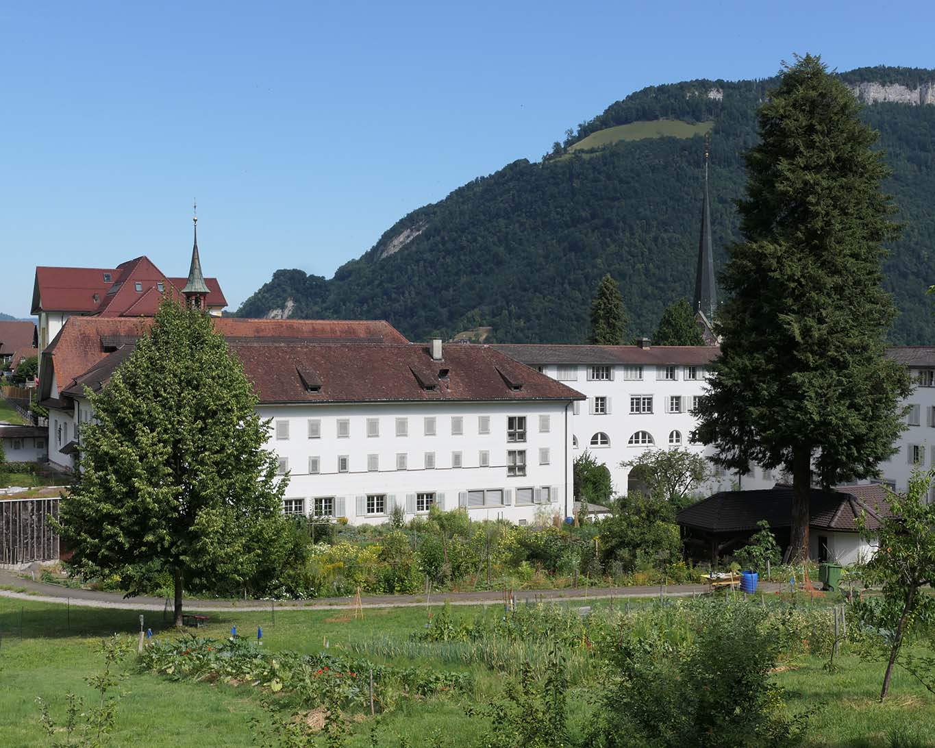 Sicht aufs Kloster St. Klara in Stans mit seinem grossen Klostergarten und den Bergen im Hintergrund bei blauem Himmel und Sonnenschein.